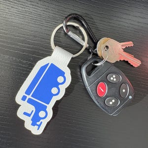 K&ouml;nnte beinhalten: Ein wei&szlig;er Leder-Schl&uuml;sselanh&auml;nger mit einem blauen LKW-Design. Der Schl&uuml;sselanh&auml;nger ist an einem silbernen Schl&uuml;sselring und einem schwarzen Karabinerhaken befestigt. Ein schwarzer Autoschl&uuml;sselanh&auml;nger mit einem roten Knopf ist ebenfalls am Schl&uuml;sselanh&auml;nger befestigt.