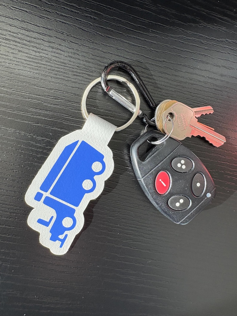 K&ouml;nnte beinhalten: Ein wei&szlig;er Leder-Schl&uuml;sselanh&auml;nger mit einem blauen LKW-Design. Der Schl&uuml;sselanh&auml;nger ist an einem silbernen Schl&uuml;sselring und einem schwarzen Karabinerhaken befestigt. Ein schwarzer Autoschl&uuml;sselanh&auml;nger mit einem roten Knopf ist ebenfalls am Schl&uuml;sselanh&auml;nger befestigt.