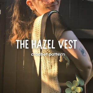 May include: A cream-colored crochet vest with a strap, featuring the text "THE HAZEL VEST crochet pattern." The vest has a textured design and is worn over a black top. A green flower pin is attached to the vest.
