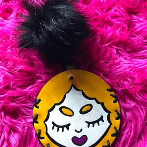 May include: A yellow leather keychain with a hand-painted face and a black fuzzy pom-pom. The face has closed eyes, a small smile, and a purple heart.
