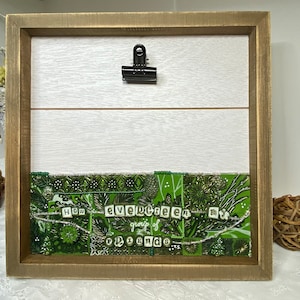 May include: A framed artwork with a green and white design. The artwork features a collage of fabric scraps with a beaded border. The text "How evergreen my group of friends is" is spelled out in white letters on the fabric.