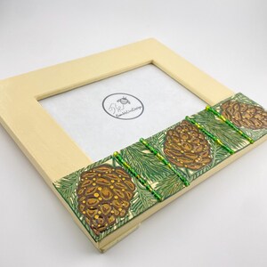 May include: A cream-colored picture frame with a green and brown pine cone design on the bottom edge. The frame has a small bee logo with the text "Round About Designs".