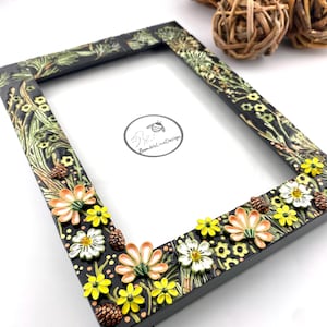May include: A rectangular black picture frame decorated with raised, colorful floral and pinecone accents. The frame features painted green foliage, yellow flowers, and peach-colored blossoms. A circular logo with a bee and the text BumbleLineDesign is visible in the center.