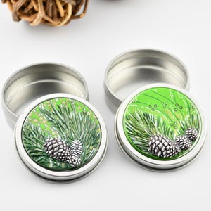 May include: Two silver round tins with green and white pine cone designs on the lids. The lids are open and the tins are empty.