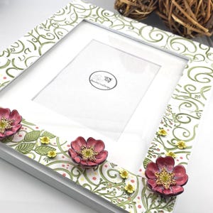 May include: A rectangular picture frame with a white border featuring a green swirling vine pattern, small yellow dots, and three dimensional pink flowers with gold centers. A circular logo with a bee and the text BumblelineDesign is visible.