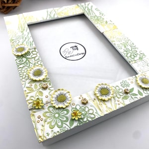 May include: White picture frame with a floral design. The frame is decorated with green and yellow flowers and leaves. The frame has a rectangular shape and is decorated with raised floral accents. The frame is approximately 8 inches by 10 inches.