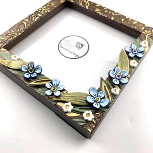 May include: A square wooden picture frame with a dark brown finish and floral embellishments. The frame features light blue flowers, green leaves, and small white flowers. The frame has the text "bumblelineldesign" in a circle.