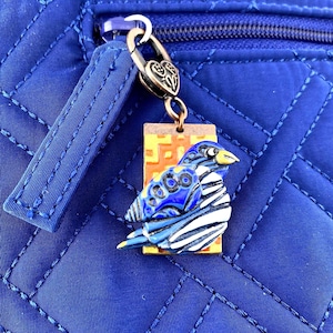 May include: A blue and white bird charm with a yellow beak hangs from a silver-colored clasp. The charm is attached to a blue quilted fabric. The bird has blue and white patterned wings and a blue body with yellow accents.
