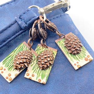 May include: Close-up of a blue leather bag with three bronze-colored pinecone charms attached. Each charm features a detailed pinecone on a rectangular base with green and orange accents. The charms are connected to the bag with a metal clasp.
