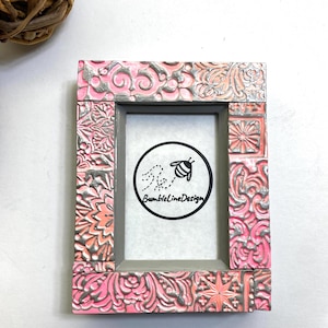 May include: A pink and gray picture frame with a floral design. The frame has a small opening for a photo and the text "BumblelineDesign" is printed on the inside.
