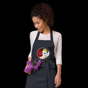 May include: A navy blue apron with a front pocket and a tie-waist. The apron features a circular graphic of a red devil and a white angel with a halo. The apron is worn by a person.