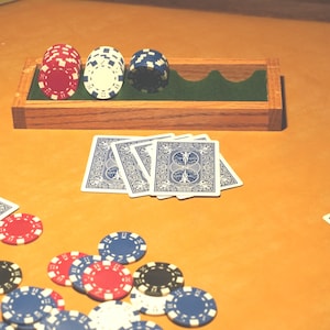 May include: A rectangular wooden tray with a green felt interior holds stacks of red, white, and blue poker chips. Below the tray, four blue playing cards with intricate white patterns are fanned out, alongside scattered red, white, and blue chips.