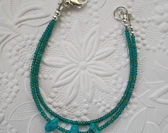May include: A double-strand bracelet featuring small, iridescent teal seed beads and three irregular, bright turquoise-colored stone beads. The bracelet is finished with silver-toned metal crimp beads, a circular jump ring, and a lobster claw clasp.