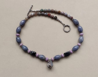 May include: A handcrafted necklace featuring a variety of beads in shades of blue, purple, black, and brown. The necklace includes oval and round beads, with a silver-tone toggle clasp. The pendant is a teardrop-shaped stone.