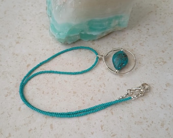 May include: A turquoise beaded necklace with a silver pendant. The pendant holds a turquoise stone. The necklace is displayed on a white and gray surface, with a large turquoise stone in the background.