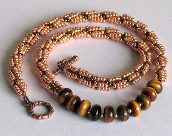 May include: A copper-colored beaded necklace with a spiral design. The necklace features a series of brown and gold tiger eye beads at the front. The clasp is a copper-colored ring and toggle.