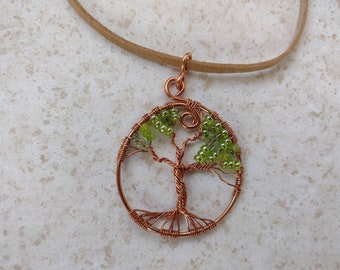 May include: A copper wire tree of life pendant with green beaded leaves, suspended from a tan suede cord. The tree design is circular, with detailed wirework forming the trunk and branches. The pendant is a handcrafted piece of jewelry.