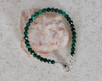 May include: A beaded bracelet featuring faceted, dark green beads. The bracelet is strung on a flexible cord and has a silver-colored clasp. The bracelet is displayed on a light-colored, rough-textured stone.