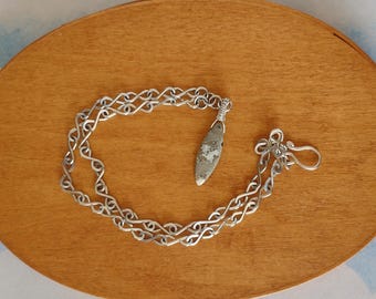 May include: A silver necklace with a woven chain design and a teardrop-shaped pendant. The pendant is a light gray color with darker gray and white markings. The necklace is displayed on a wooden oval tray.