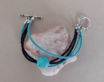 May include: A multi-strand bracelet featuring alternating rows of small black and turquoise seed beads. A single larger, round turquoise bead is centered on the strands. The bracelet is secured with a silver-toned metal toggle clasp.