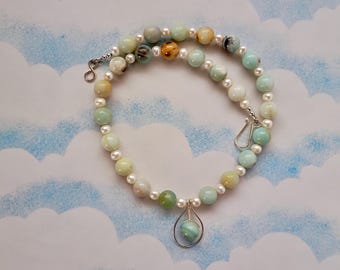 May include: A beaded necklace featuring a mix of round, light green, and cream-colored beads, interspersed with small white pearls. A teardrop-shaped pendant with a light green stone hangs from the center. The necklace has a silver clasp.