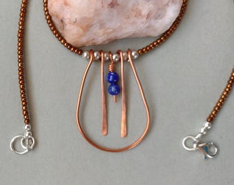 May include: A handcrafted copper pendant necklace with a teardrop design. The necklace features a beaded chain, silver accents, and two blue lapis lazuli beads. The pendant is made of copper wire and has a unique, artistic look.