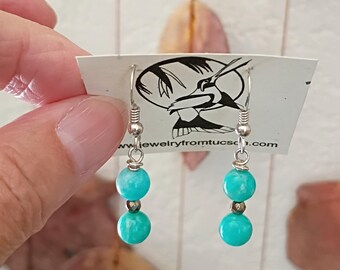 May include: A pair of turquoise bead earrings with silver-colored hooks. Each earring features two turquoise beads and a smaller silver bead. The earrings are displayed on a white tag with a black and white hummingbird design and the website jewelryfromtucson.com.