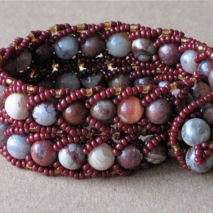 May include: A multi-strand bracelet constructed from round, variegated earth-toned stone beads in shades of brown, cream, and gray. The beads are intricately woven together with small, deep red seed beads and accented with tiny, translucent gold beads.