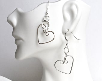 May include: Silver heart-shaped earrings with a wire design. The earrings feature a hook closure and are connected by small circular links. The heart shape has a curled detail at the bottom. The earrings are displayed on a white mannequin.