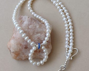 May include: A white pearl necklace with a double strand design. The necklace features a small blue accent and a silver clasp. The pearls are round and evenly sized. The necklace is displayed on a light pink rock against a neutral background.