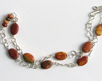 May include: A silver chain necklace with oval-shaped, reddish-brown and yellow-orange jasper beads. The necklace has a double strand design with a hook clasp. The beads are connected by silver chain links.