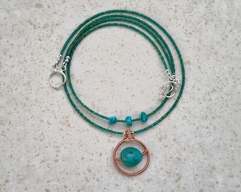 May include: A necklace featuring a long strand of small, dark green beads with three small turquoise stones and a central pendant. The pendant consists of a round, copper-colored wire frame holding a single, round, speckled turquoise stone.
