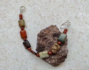May include: A brown, green, and red stone bracelet with a silver clasp. The bracelet is made of rectangular stones and is lying on a rock.
