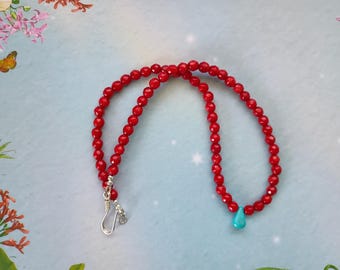 May include: A necklace composed of small, faceted red beads arranged in a circular pattern. A single, smooth, teardrop-shaped turquoise bead hangs from the strand. The necklace features a silver-toned metal hook clasp at the ends.