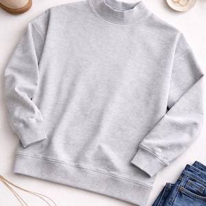 Peut inclure: Un sweat-shirt gris clair à col montant avec des poignets et un ourlet côtelés. Le sweat-shirt est associé à un jean bleu, des bottes beiges et des bijoux dorés. Une tenue décontractée.
