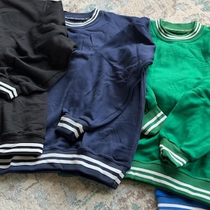 May include: A collection of crew neck sweatshirts in black, navy blue, green, and blue. Each sweatshirt has white stripes on the cuffs, collar, and hem. The sweatshirts are made of a soft material.
