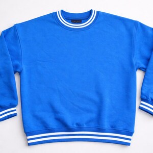 May include: A vibrant blue sweatshirt with white stripes on the collar, cuffs, and hem. The long-sleeved top is made of a soft material and has a classic, sporty design. This casual garment is suitable for everyday wear.