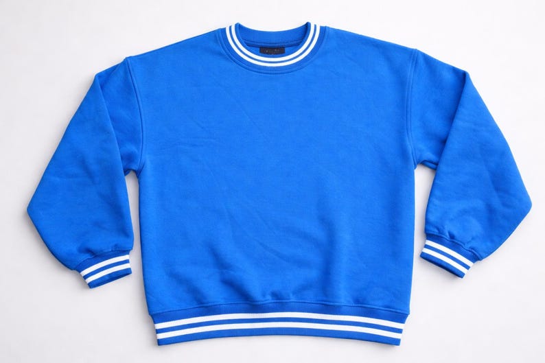 May include: A vibrant blue sweatshirt with white stripes on the collar, cuffs, and hem. The long-sleeved top is made of a soft material and has a classic, sporty design. This casual garment is suitable for everyday wear.