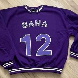 May include: A purple sweatshirt with the name "SANA" and the number "12" in white lettering. The sweatshirt has white stripes on the collar, cuffs, and hem. The sleeves are long and the overall style is casual.
