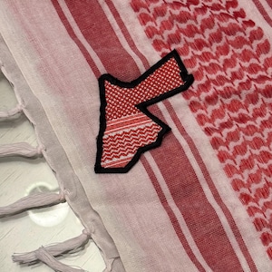 Könnte beinhalten: Ein rot-weißes Keffiyeh-Tuch mit einem schwarz umrandeten Aufnäher in Form von Jordanien. Das Tuch hat ein gestreiftes und gemustertes Design mit Quasten am Rand. Der Aufnäher ist rot und weiß.