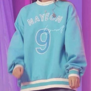 May include: A light blue sweatshirt with white trim and the name "NAYEON" in white block letters. Below the name is the number "9" in white, and a cursive signature. The sweatshirt has long sleeves and a crew neck.
