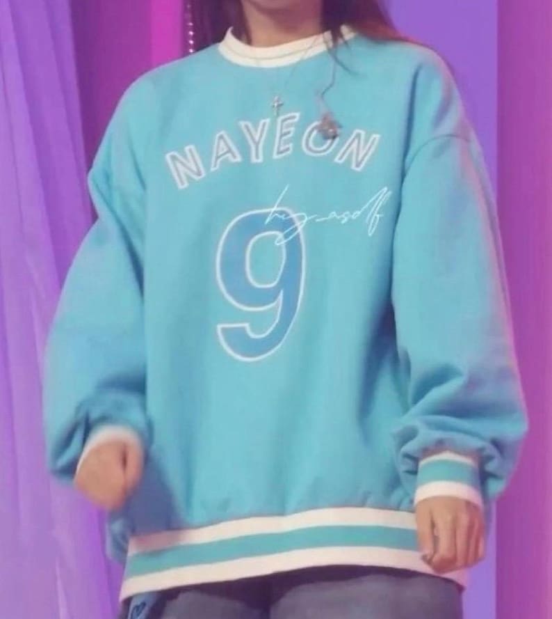 May include: A light blue sweatshirt with white trim and the name "NAYEON" in white block letters. Below the name is the number "9" in white, and a cursive signature. The sweatshirt has long sleeves and a crew neck.
