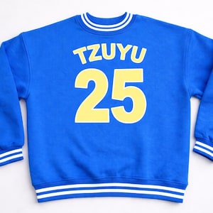 May include: A royal blue sweatshirt with the name "TZUYU" in yellow block letters above the number "25". The sweatshirt has white and yellow striped details on the collar, cuffs, and hem.