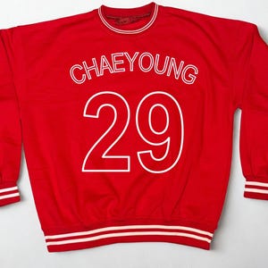 May include: A red sweatshirt with white lettering and trim. The word "CHAEYOUNG" is arched across the top, with the number "29" below. The cuffs and collar have white stripes.