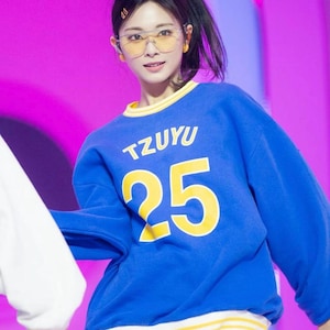 May include: A person wearing a blue sweatshirt with the word "TZUYU" and the number "25" in yellow. The sweatshirt has yellow and white trim. The person is also wearing yellow-tinted glasses.