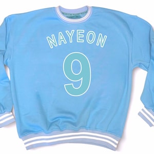May include: Light blue sweatshirt with the name "NAYEON" in white letters above a large number "9" in teal. The cuffs and collar have white and light blue stripes. The sweatshirt is a casual style.