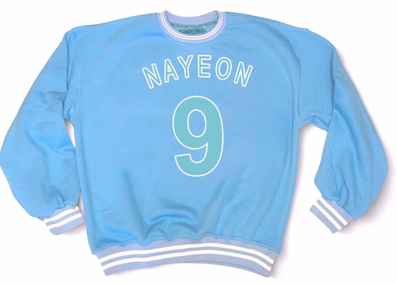 May include: Light blue sweatshirt with the name "NAYEON" in white letters above a large number "9" in teal. The cuffs and collar have white and light blue stripes. The sweatshirt is a casual style.