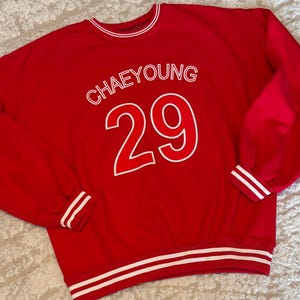 May include: A red sweatshirt with white accents. The word "CHAEYOUNG" is printed above the number "29" in white. White stripes are on the cuffs, collar, and hem.