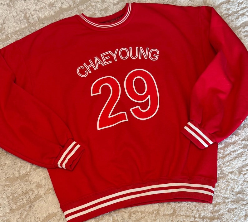 May include: A red sweatshirt with white accents. The word "CHAEYOUNG" is printed above the number "29" in white. White stripes are on the cuffs, collar, and hem.