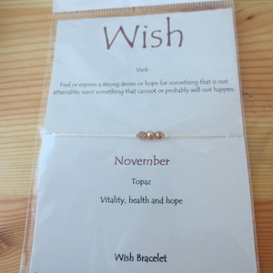 May include: A white string bracelet with a small topaz gemstone charm. The card reads "Wish" and "November Topaz Vitality, health and hope". It also says "Wish Bracelet Close your eyes and make a wish. Tie the bracelet around your wrist or ankle. When your bracelet naturally breaks in two, your wish is ready to come true..."